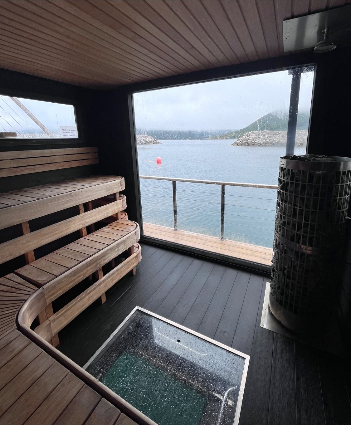Rainy dusk view of the fjord from inside the sauna, with the wood-fired stove in frame