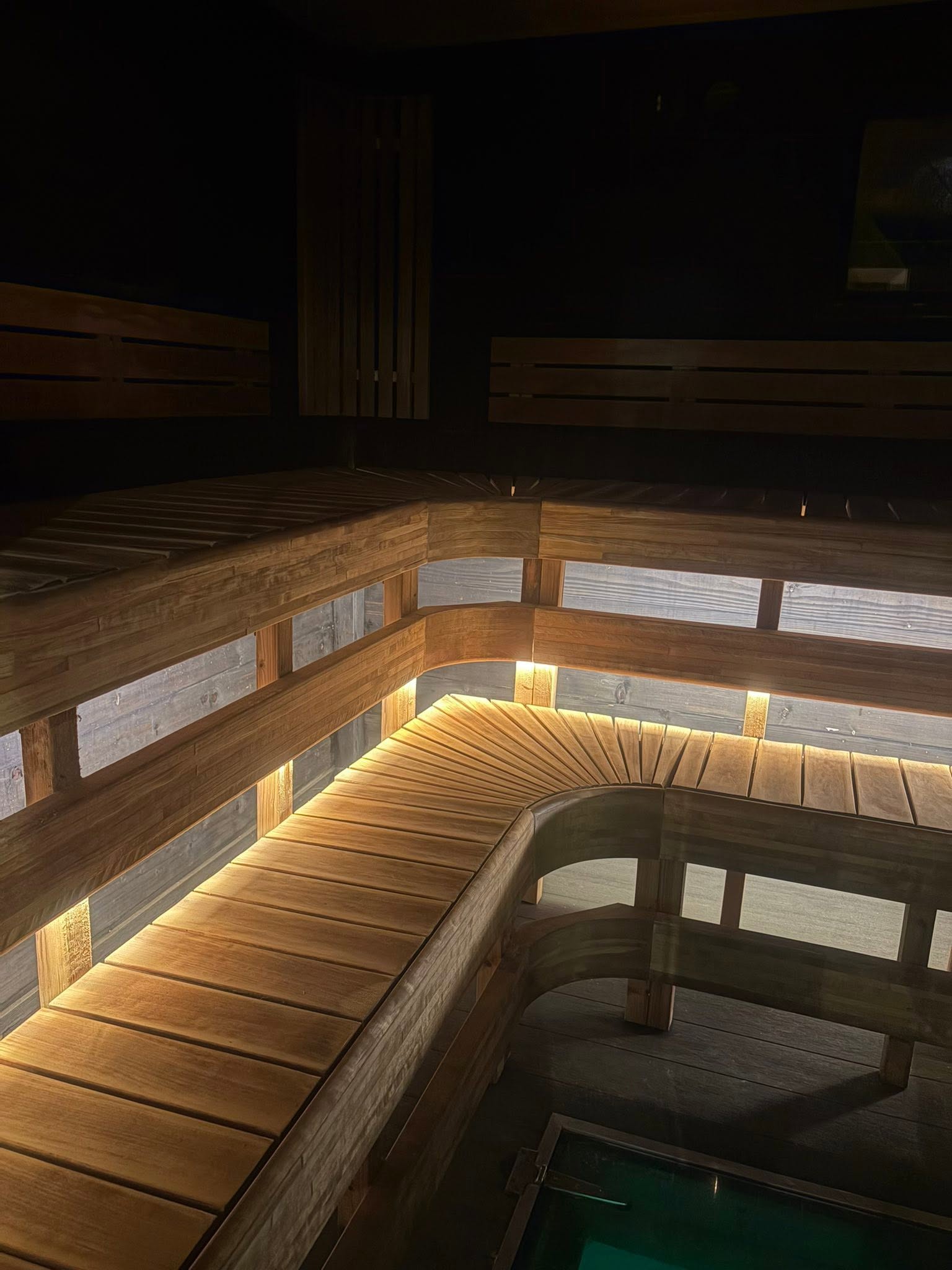 Curved sauna benches lit by warm LED strips at night, glass plunge pool in the floor