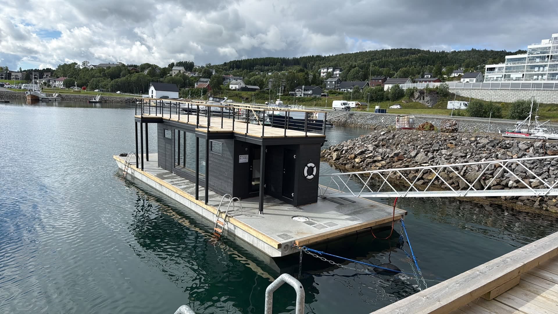 Damperiet's floating sauna moored in Olderhamna harbour, Finnsnes, with a rooftop deck and mountains behind